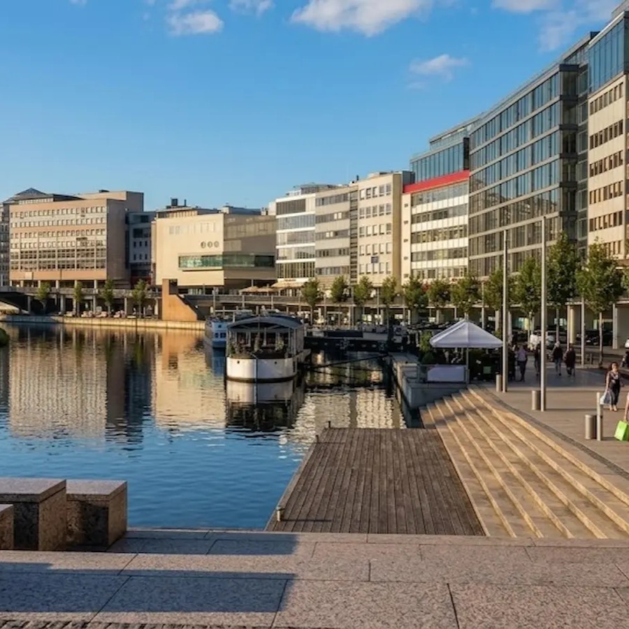 Blick auf die Saarpromenade in Saarbrücken mit Stadtgebäuden, Wasser und Fußgängern.